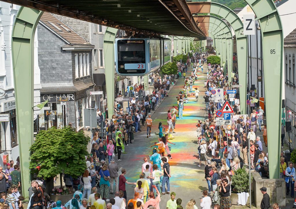 Viele Menschen bewegen sich unter der Wuppertaler Schwebebahn über eine farbig gestaltete Straße, die von Autos befreit wurde. Die Szene zeigt eine künstlerische Performance im öffentlichen Raum mit zahlreichen Beteiligten und Zuschauer*innen.