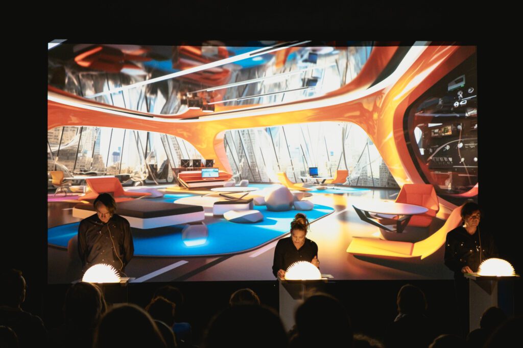Drei Mitwirkende der performativen Film-Lesung 2052 – Nachrichten aus der Zukunft stehen auf einer Bühne vor einer großformatigen Projektion einer futuristischen Innenarchitektur-Szene.