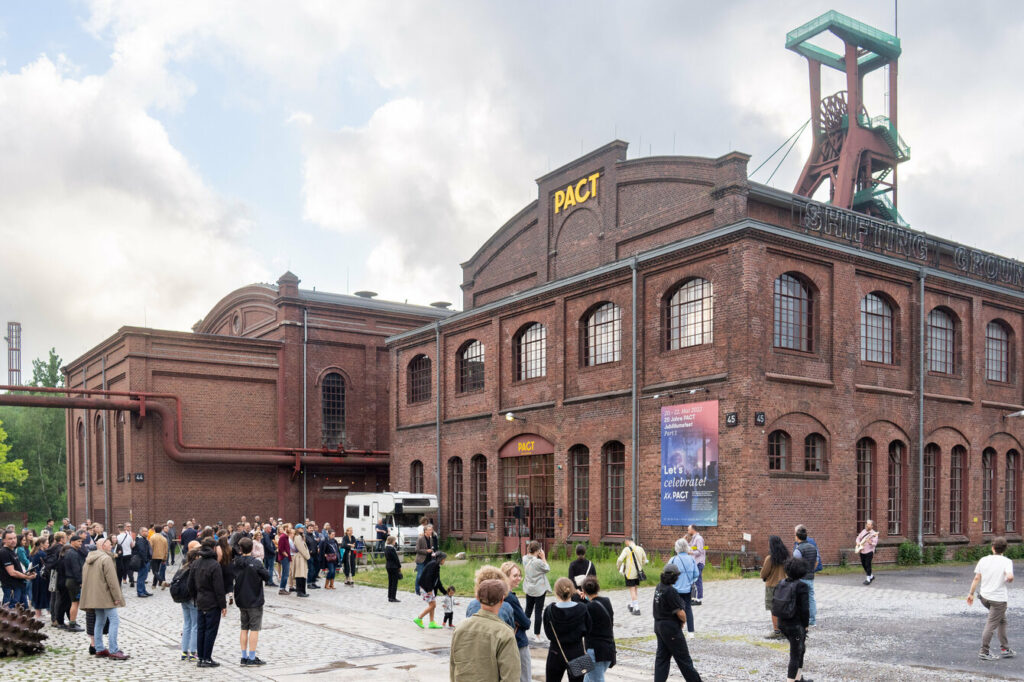 Außenansicht des Choreographischen Zentrums NRW (PACT) aus rotem Backstein mit einem Förderturm im Hintergrund bei einer Veranstaltung. Zahlreiche Besucher*innen versammeln sich auf dem gepflasterten Vorplatz. An der Spitze des historischen Industriegebäudes ist ein großes, gelbes PACT-Logo angebracht und an der Wand hängt ein blaues Veranstaltungsplakat mit der Aufschrift „Let’s celebrate! / 20 Jahre PACT“.