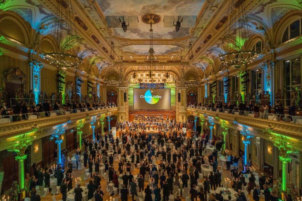 Zahlreiche Menschen stehen an festlich gedeckten Tischen im Saal und auf den Balkonen der Historischen Stadthalle Wuppertal. Sie sind zur Bühne gewandt und applaudieren einem Orchester bei einer Veranstaltung der Stiftung.