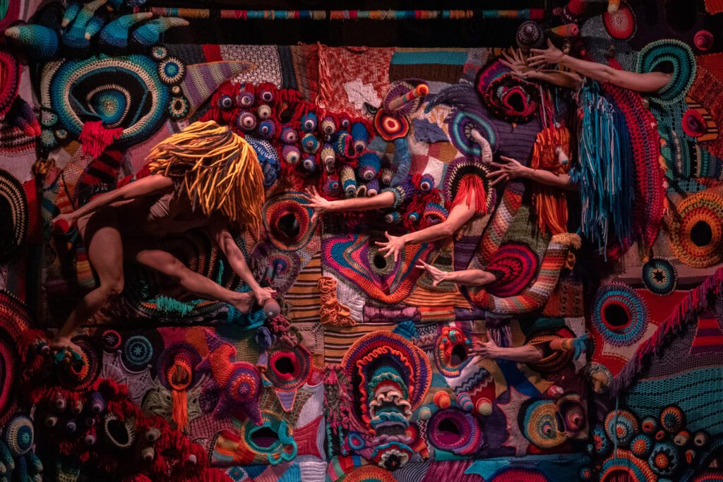 Mehrere Performende bewegen sich vor und zwischen einer gro&szlig;fl&auml;chigen, farbenfrohen Installation aus geh&auml;kelten und gestrickten Textilien. Arme und Beine ragen aus der textilen Struktur hervor und verschmelzen mit ihr zu einer gemeinsamen B&uuml;hnenszene.