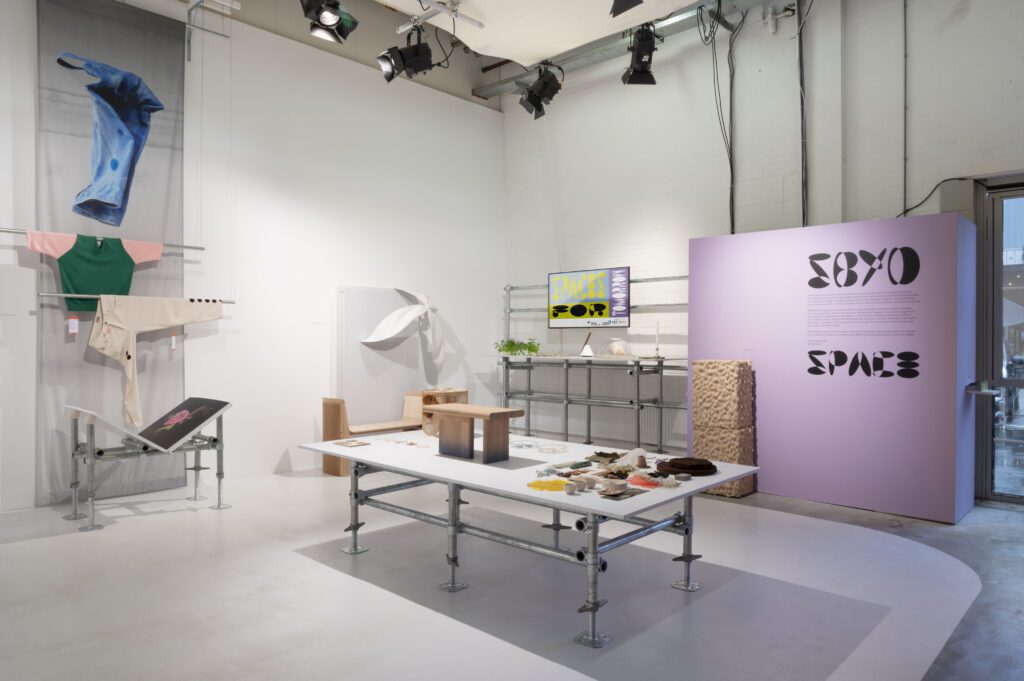Heller Ausstellungsraum mit Designobjekten und Materialproben: Ein zentraler Tisch zeigt verschiedene Exponate, daneben hängen Kleidungsstücke an einer Metallkonstruktion. Im Hintergrund sind Sitzmöbel, ein Bildschirm und weitere Ausstellungselemente zu sehen.
