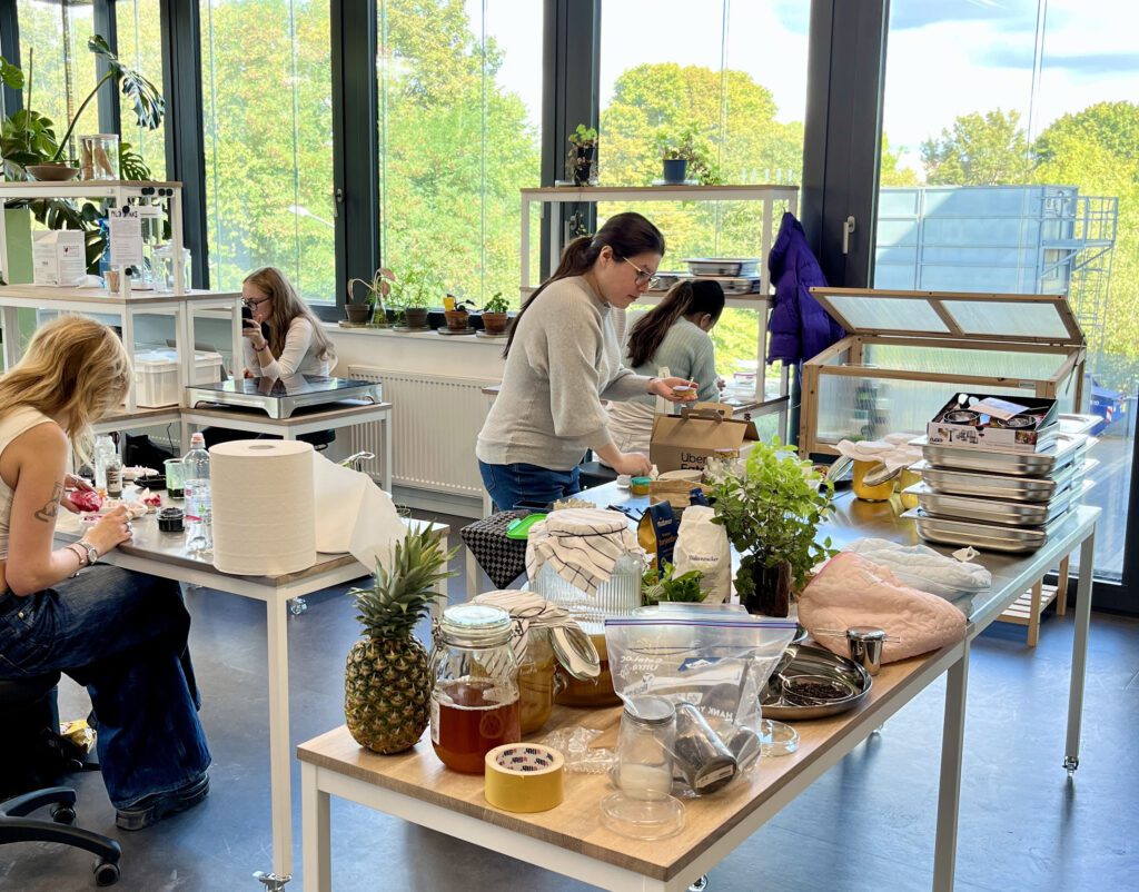 Arbeit im BioLab der Folkwang Universität der Künste: Vier Personen arbeiten in einem hellen Laborraum an verschiedenen Tischen mit Pflanzen, Lebensmitteln und weiteren Materialien. Auf den Arbeitsflächen stehen unter anderem eine Ananas, Gläser mit fermentierten Lebensmitteln, Gewürze, Kräuter, Waagen und Schalen. Im Hintergrund sind Regale, ein kleines Gewächshaus, viele Pflanzen und große Fenster mit Blick ins Grüne zu sehen.