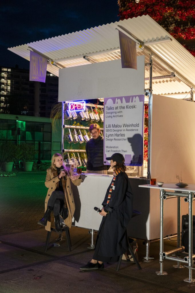 Podiumsgespräch an einem temporären Kiosk: Zwei Personen sprechen mit Mikrofonen miteinander, während eine dritte Person hinter dem Tresen steht. Ein Plakat mit dem Titel “Talks at the Kiosk: Designing with Living Archives” ist am Kiosk zu sehen.