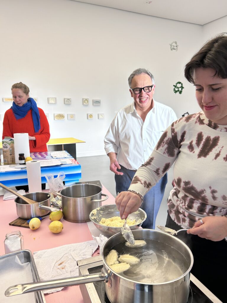 The artist and two other people are preparing food together at a table in an exhibition space. In the foreground, one person is using a spoon to add pieces of dough to a large pot; on the table are additional pots, a bowl of dough, lemons, and kitchen utensils. 