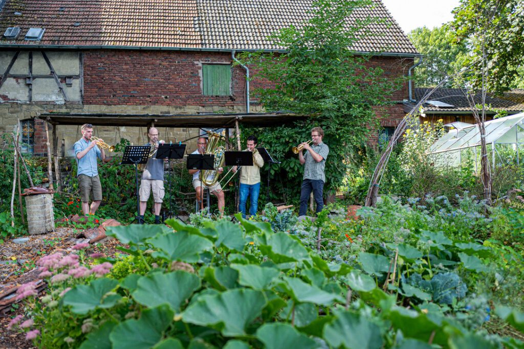 F&uuml;nf Mitglieder des Orchesters spielen ein St&uuml;ck in einem Garten zwischen verschiedenen Gr&uuml;npflanzen.
