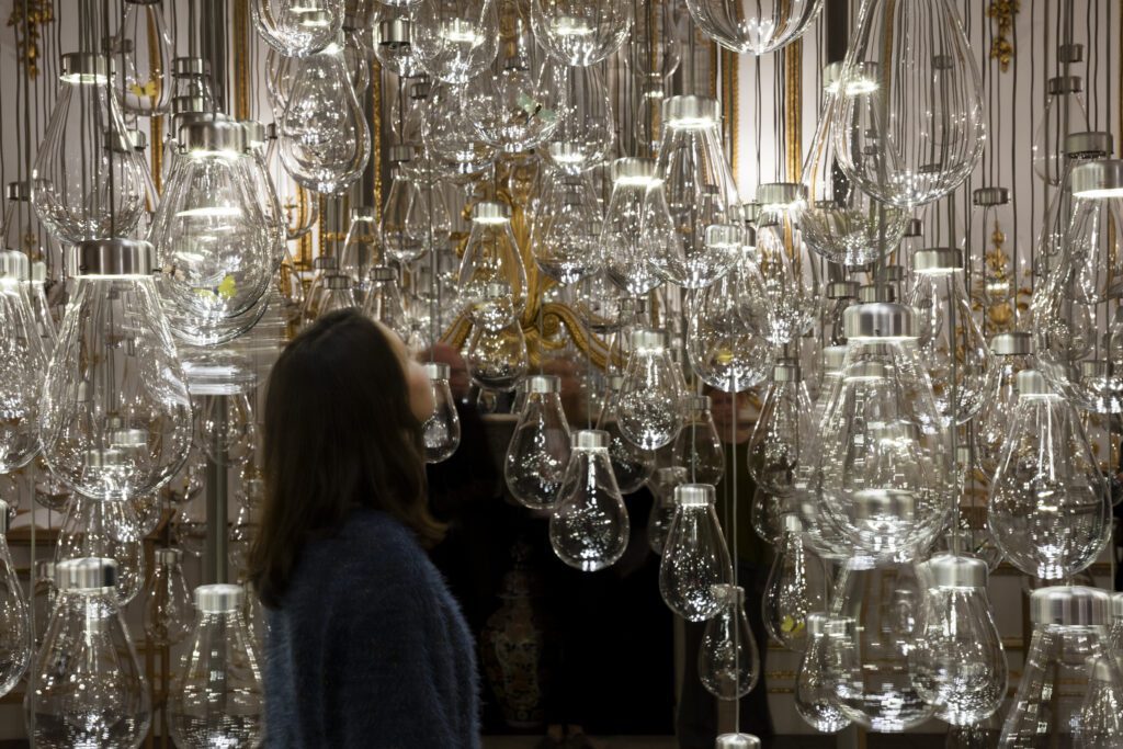 Rauminstallation mit einer F&uuml;lle von von der Decke h&auml;ngenden Glaskugeln. Die Kugeln erinnern an Gl&uuml;hbirnen und enthalten Insektenfiguren in realit&auml;tsnaher Gr&ouml;&szlig;e. Eine Museumsbesucherin betrachtet die Installation aus der N&auml;he.