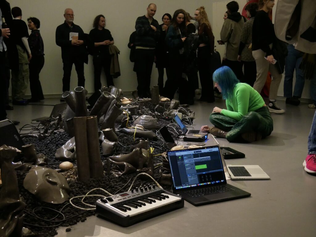 Die K&uuml;nstlerin (Maria Wildeis) sitzt vor mehreren Laptops und elektronischen Instrumenten neben einer keramischen Installation, w&auml;hrend Besucher*innen die Performance beobachten.