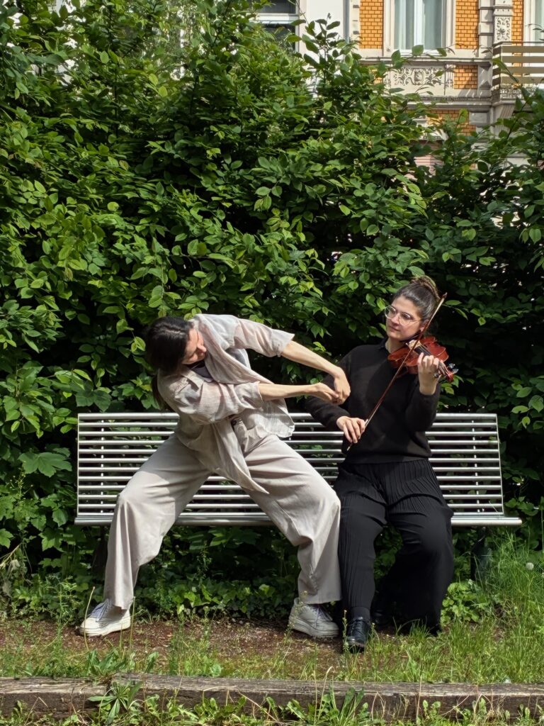 Eine musikalische Tanzperformance im Grünen: Zwei Frauen auf einer Parkbank vor einer dichten grünen Hecke – eine der beiden spielt Geige, die andere tanzt mit ausdrucksstarker Pose.