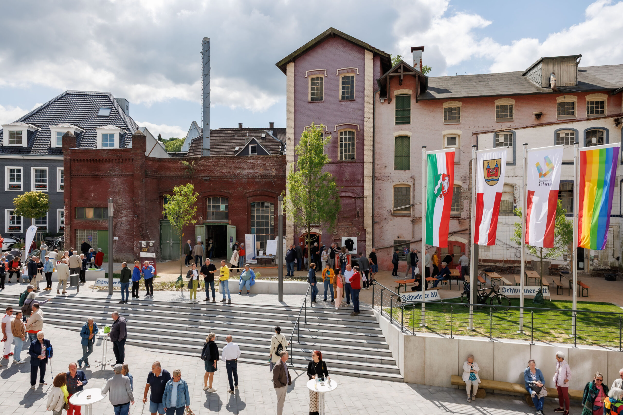 A bustling square filled with people in front of the historic factory buildings of the former brewery. The large gates to the old brewery are open, and visitors are coming and going. Next to them, four large, colorful flags flutter in the wind, including the flag of the state of North Rhine-Westphalia and a rainbow flag symbolizing the LGBTQ+ movement.