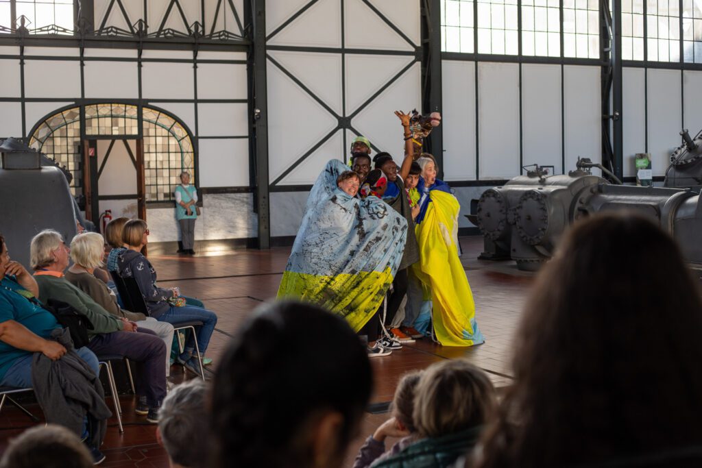 Several performers in an indoor space interact in front of an audience with textile materials and fabrics that are integrated into a shared performative action. The performers are crowded together and enveloped by the fabric as they move, while simultaneously working with the fabric, which forms the central focus of the scene.   