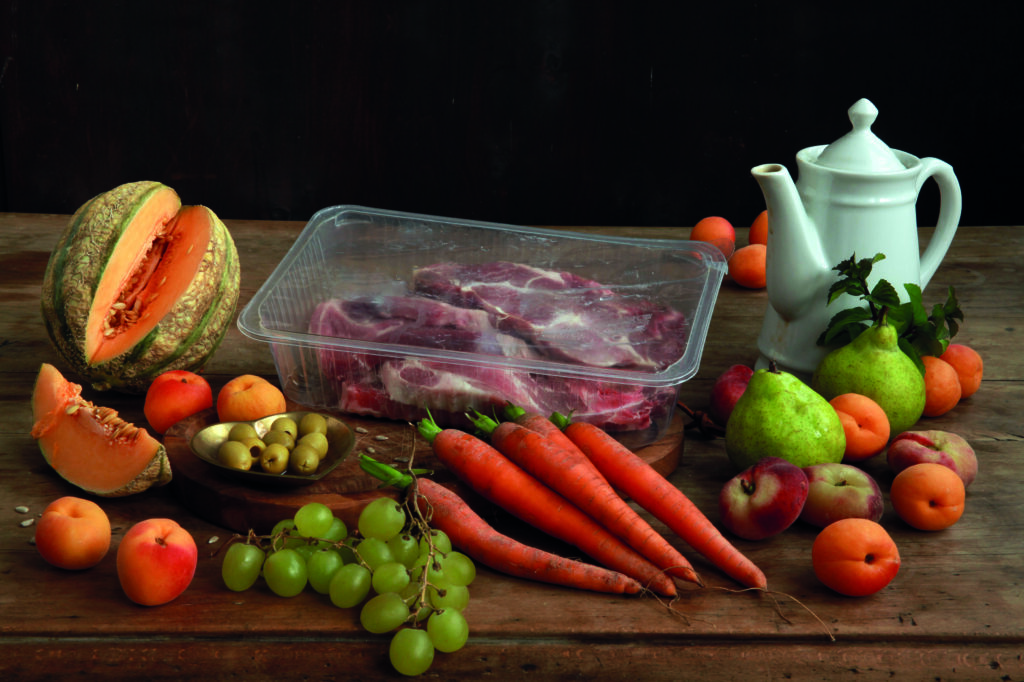 A photographic still life featuring fruits and vegetables on a set table. A porcelain pitcher stands next to it. Among the produce, meat products wrapped in plastic wrap are arranged against a dark background.