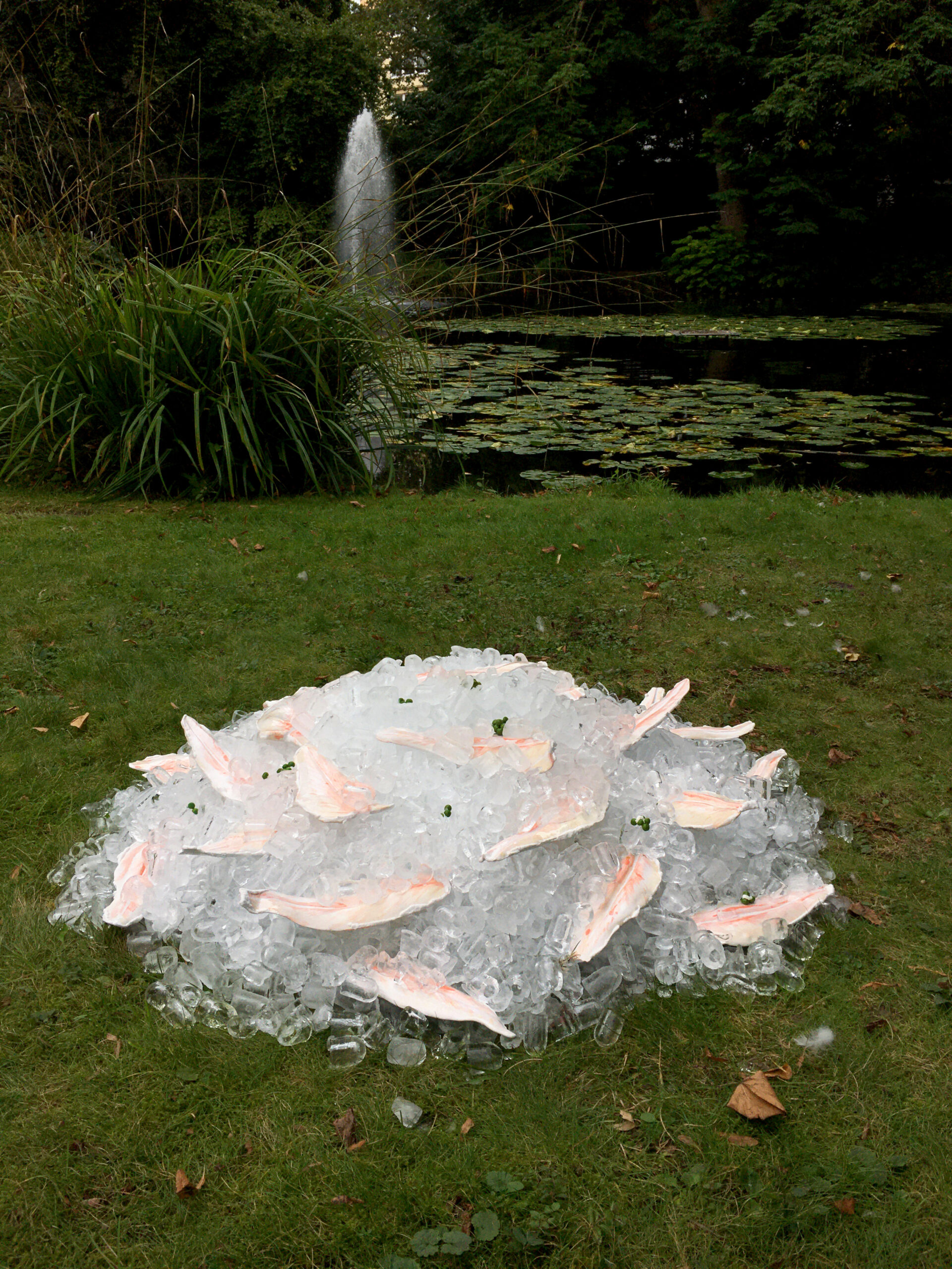 Installation &bdquo;Pangasius&ldquo; von Katharina Maderthaner: Keramikobjekte in Form von Fischfilets und Erbsen liegen auf einem H&uuml;gel aus Eisw&uuml;rfeln auf einer Wiese vor einem Teich.