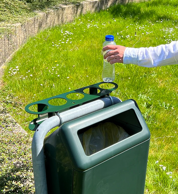 A person places a returnable bottle into a green return bottle ring with several round bottle openings next to a public trash can in a park.