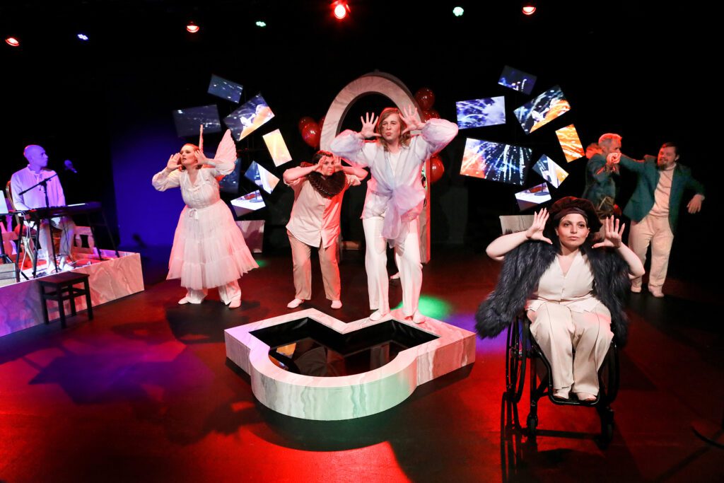 Several performers are standing and moving around on a theater stage as part of a group performance. In the foreground, a person in a wheelchair is sitting with their hands raised; other people are striking similar poses. Monitors and a keyboard can be seen on the stage in the background.  