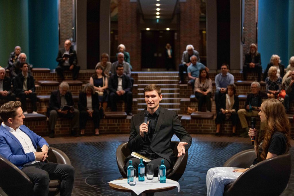 Podiumsgespräch im Foyer der Tonhalle in Düsseldorf vor Publikum. Drei Personen sitzen mit Mikrofonen auf einer Bühne und diskutieren im Rahmen der Konzertreihe Green Monday.