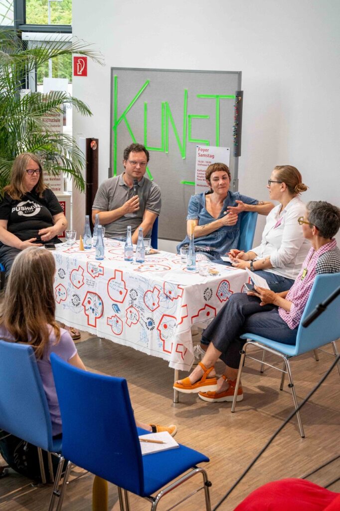 Personen sitzen an einem Tisch und sprechen miteinander. Auf der Tischdecke sind handschriftliche Notizen aus der gemeinsamen Arbeit zu sehen.