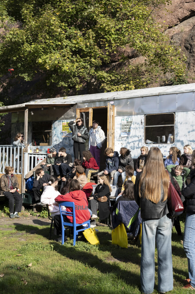 Mehrere Menschen sitzen und stehen vor einem zu einem Aufenthaltsraum umgebauten Eisenbahnwaggon bei einer Summer-School-Veranstaltung der Kunsthochschule Halle.
