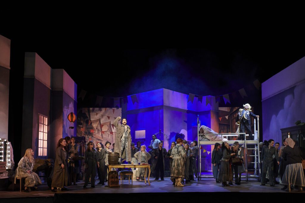 Mehrere Darstellende spielen eine Szene auf einer Theaterb&uuml;hne vor einem mehrteiligen B&uuml;hnenbild mit Podesten und beweglichen B&uuml;hnenelementen.