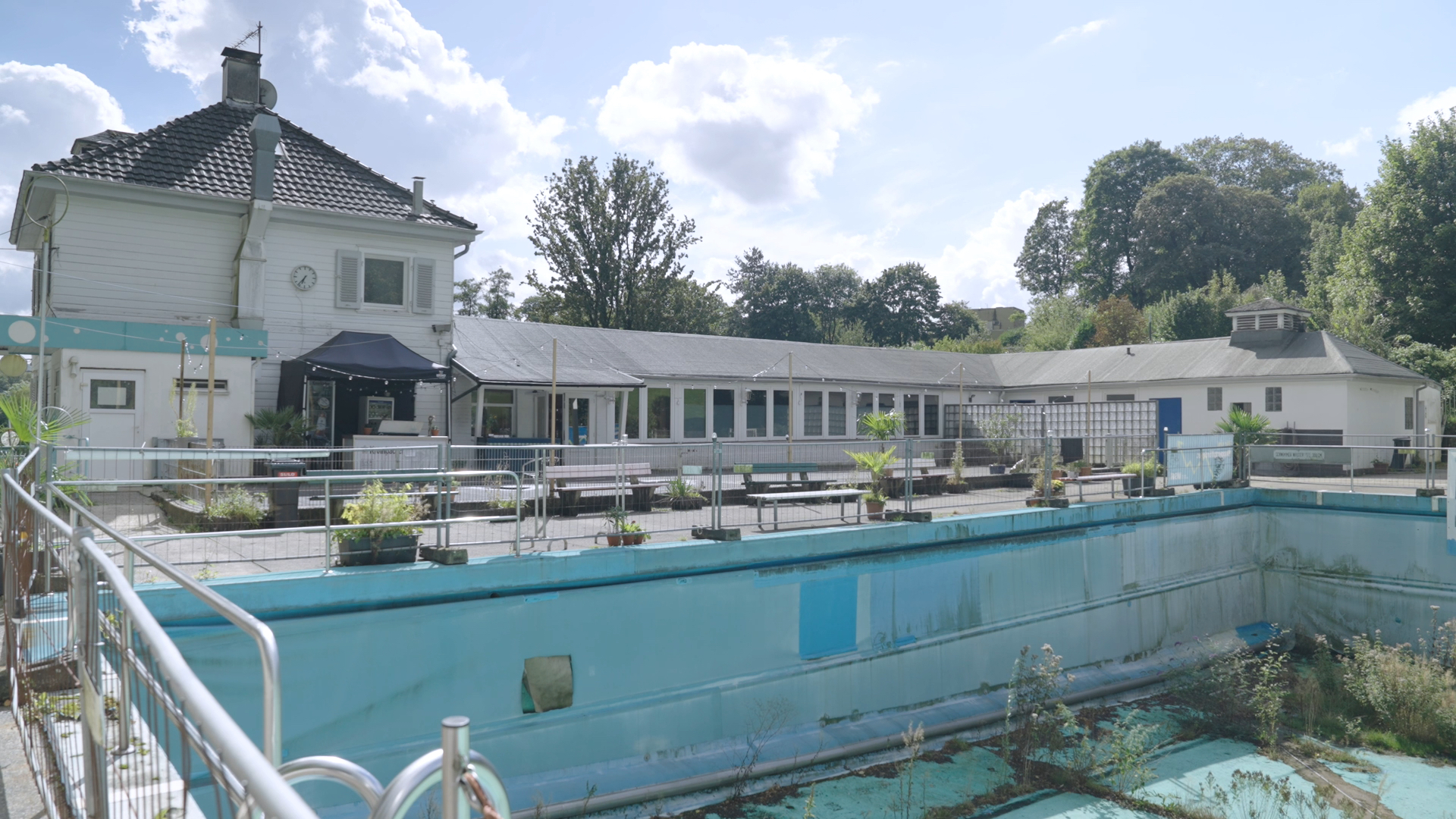 Blick auf ein leeres, hellblau gestrichenes Schwimmbecken, in dem einige Pflanzen wachsen und welches von einem Bauzaun umrandet ist. Im Hintergrund stehen flache, weiße Gebäude mit einer Terrasse, Bänken und Topfpflanzen.