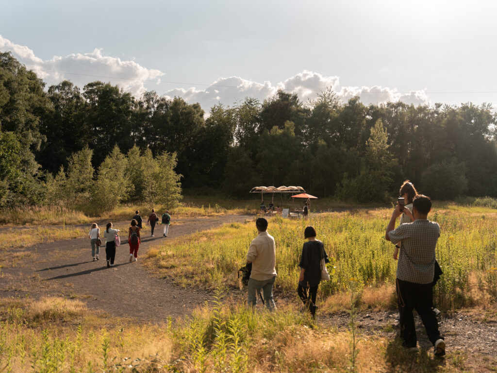 Besucher*innen bewegen sich über die begrünte Halde der Zeche Zollverein zu einer Station des Shifting Grounds Festivals im Freien.