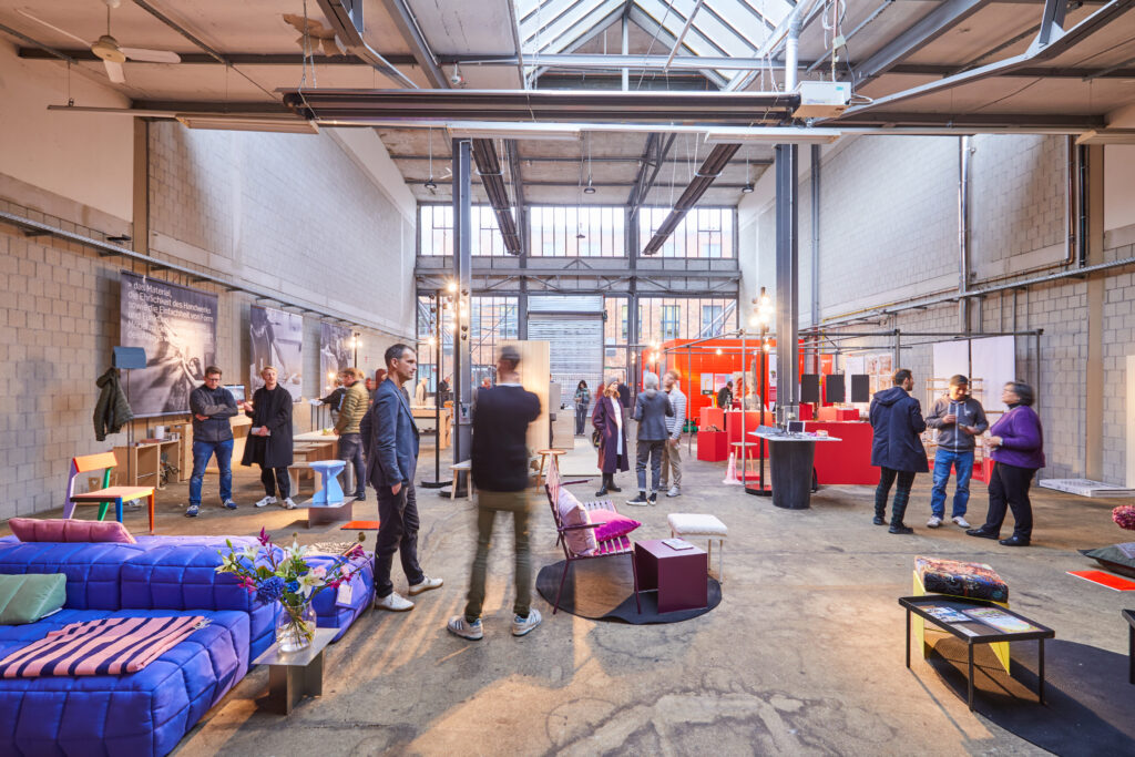 A design exhibition in a large, light-filled industrial hall with skylights and exposed walls. Modern upholstered furniture takes center stage, while in the background, exhibition booths and visitors engaged in conversation can be seen.