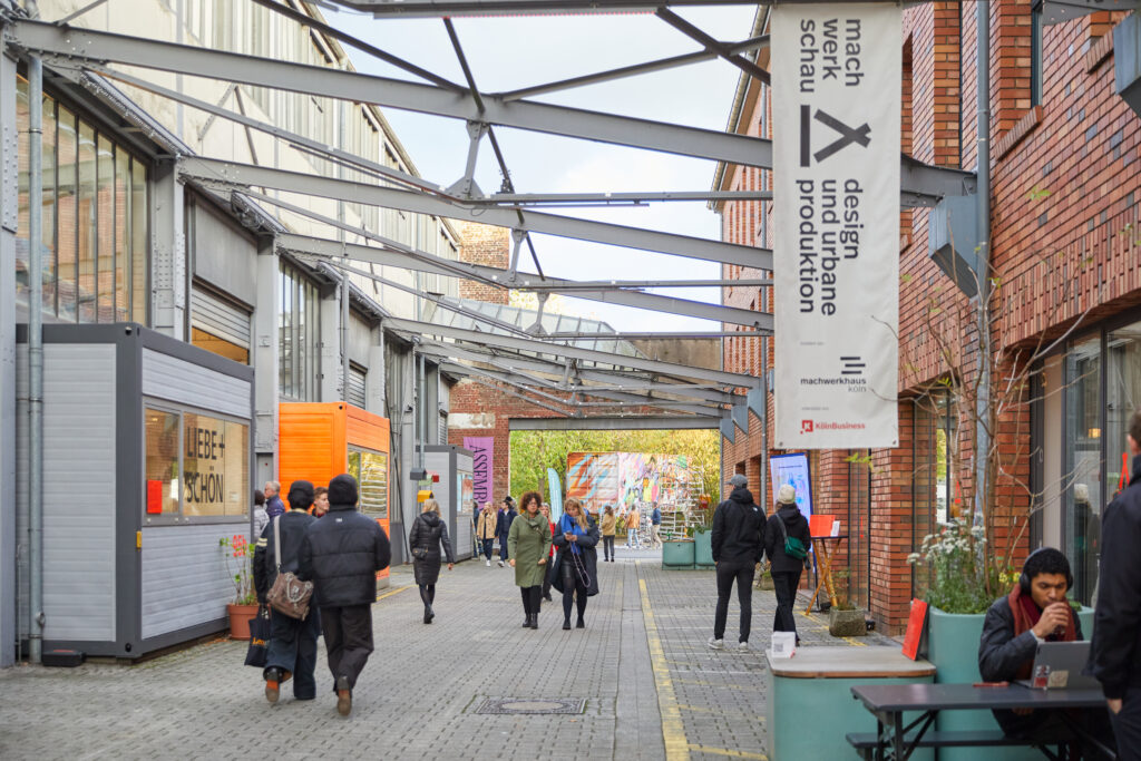 Outside the machwerkhaus köln: A paved courtyard between brick industrial buildings with a few visitors. In the foreground hangs a large white banner reading “machwerkschau – design and urban production,” along with the logos of machwerkhaus köln and KölnBusiness.