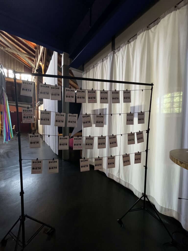A rack with hanging cards labeled “Wanted” and “Offered.” It serves as an analog bulletin board where people can share offers, requests, or interests and thus make new connections.